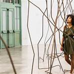 A young adult with brown textured curly hair wearing a dark green dress walking is walking through an art installation with multiple thin bent metal poles.